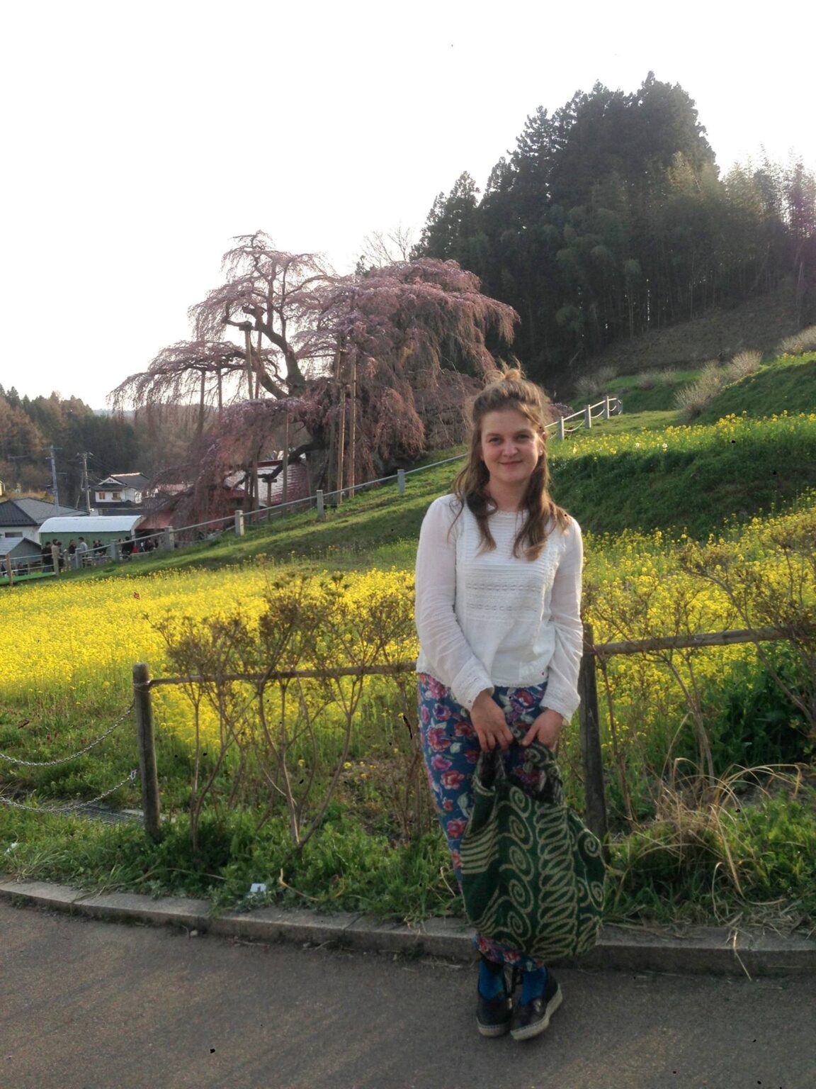 Full body portrait of ceramic artist Timi Lantos leaning against a fence in a garden with a flowering tree behind her